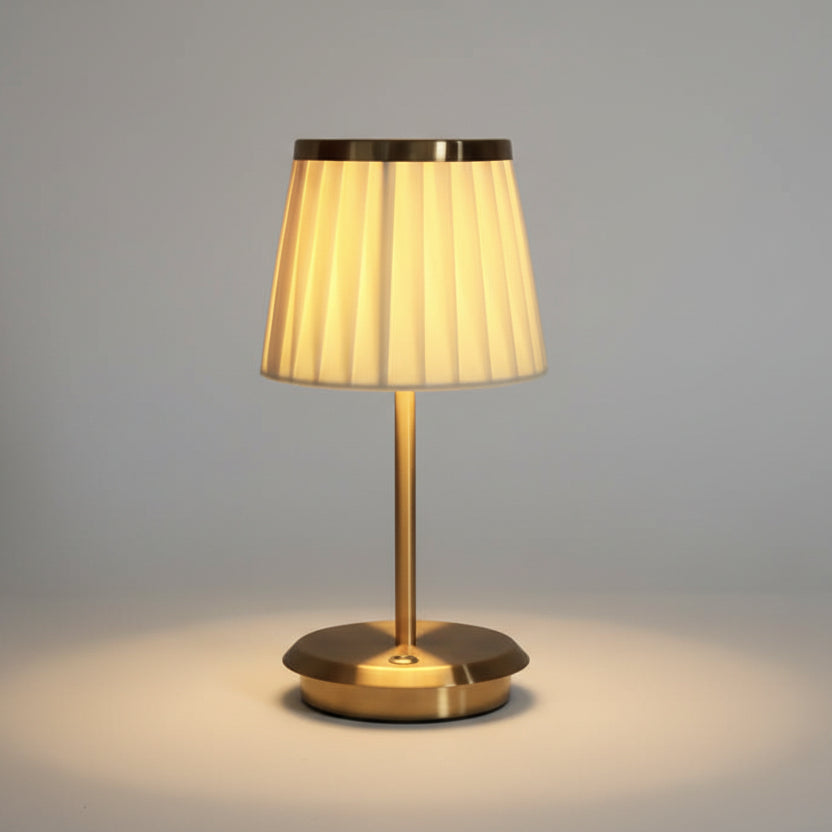 Lampe à poser moderne avec abat-jour plissé de couleur ivoire et pied fin en métal doré, offrant un éclairage chaleureux et tamisé pour une chambre ou un salon contemporain.